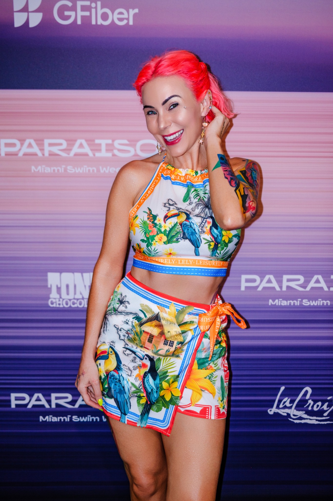 Chelsea Patricia with vivid coral-pink hair, smiling on a Miami Swim Week step-and-repeat backdrop in a colorful printed two-piece set